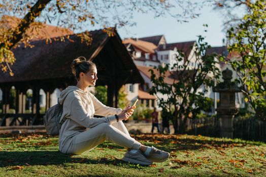 Young fashionable teenage girl with smartphone in park in autumn sitting at smiling. Trendy young woman in fall in park texting. Retouched, vibrant colors. Beautiful blonde teenage girl wearing casual modern autumn outfit sitting in park in autumn. Retouched, vibrant colors, brownish tones.