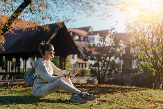 Young fashionable teenage girl with smartphone in park in autumn sitting at smiling. Trendy young woman in fall in park texting. Retouched, vibrant colors. Beautiful blonde teenage girl wearing casual modern autumn outfit sitting in park in autumn. Retouched, vibrant colors, brownish tones.