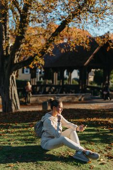 Young fashionable teenage girl with smartphone in park in autumn sitting at smiling. Trendy young woman in fall in park texting. Retouched, vibrant colors. Beautiful blonde teenage girl wearing casual modern autumn outfit sitting in park in autumn. Retouched, vibrant colors, brownish tones.