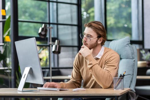 Thinking blond businessman at work inside modern green loft office, young man in casual clothes seriously ponders decision and completes task.