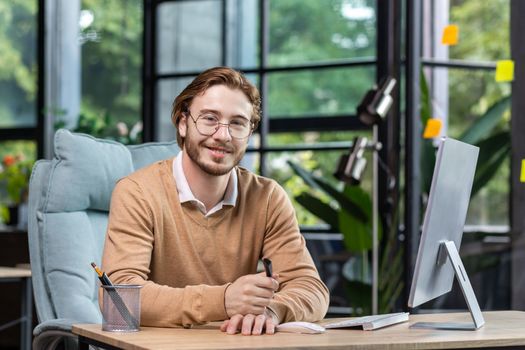 Portrait of successful smiling programmer inside modern green loft office, blond man smiling and looking at camera, businessman in sweater and casual shirt close up working with computer.