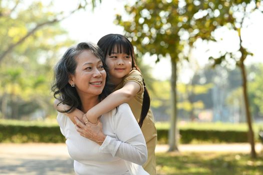 Image of smiling idle age grandmother giving piggyback ride to cute little granddaughter during walking on in the park.