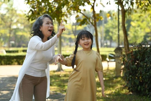 Joyful little girl and grandmother walking in public park surrounded by green trees at sunlight morning. Family, generation concept.