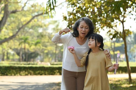 Happy middle age grandmother and lovely granddaughter blowing soap bubbles, spending leisure weekend time together at outdoor.