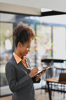 Portrait of American African female employee standing in modern office interior and using digital tablet.