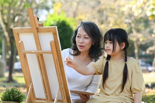 Lovely little girl and grandmother drawing in summer park, spending leisure weekend at outdoor together.