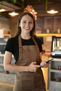 Portrait of young female barista standing front of counter in modern cafe and smiling to camera.