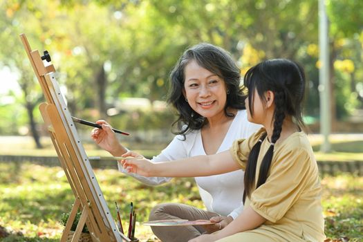 Lovely little girl and grandmother drawing in summer park, spending leisure weekend at outdoor together.