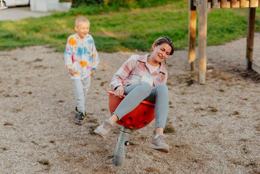 Mother and son having fun on a swing. Motherhood and childhood concept. Sunny summer day. Beautiful family is having fun outside. Parents with children riding on a swing. Mom is playing with her little son on a terrace with swings. A mother woman swings on a swing in the park. Mom and child are on a swing in nature