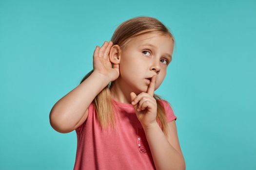 Close-up shot of a nice blonde little lady with two ponytails on her head, in a pink dress, acting like listening to someone while posing against a blue background with copy space. Concept of a joyful childhood.