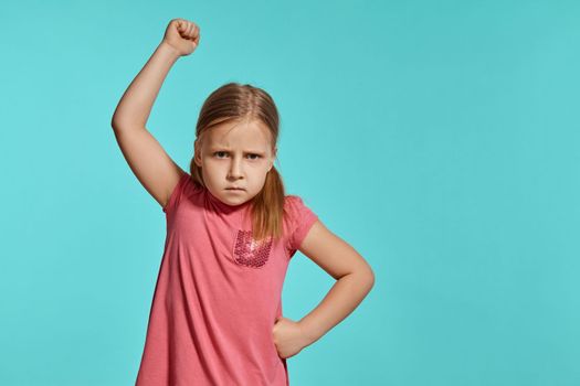 Close-up shot of a cute blonde little lady with two ponytails on her head, in a pink dress, being mad about something and raising her hand up while posing against a blue background with copy space. Concept of a joyful childhood.