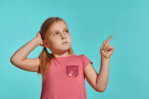 Close-up shot of a pretty blonde little female with two ponytails on her head, in a pink dress, pointing on something and looking up while posing against a blue background with copy space. Concept of a joyful childhood.