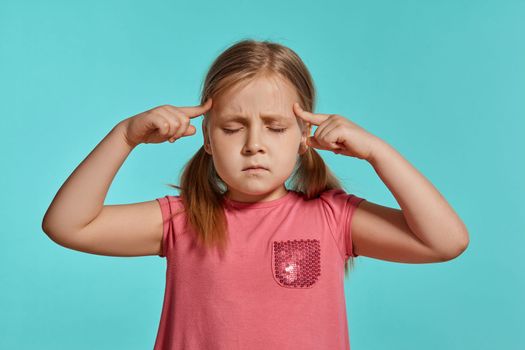 Close-up shot of a pretty blonde kid with two ponytails on her head, in a pink dress, thinking about something with closed eyes while posing against a blue background with copy space. Concept of a joyful childhood.