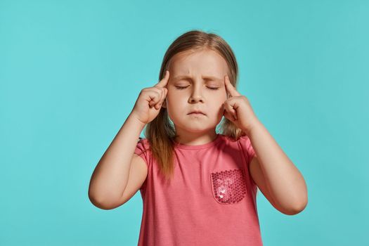 Close-up shot of pretty blonde child with two ponytails on her head, in a pink dress, thinking about something while posing against a blue background with copy space. Concept of a joyful childhood.