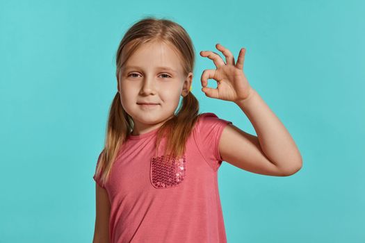 Close-up shot of a beautiful blonde little female with two ponytails on her head, in a pink dress, showing ok sign against a blue background with copy space. Concept of a joyful childhood.