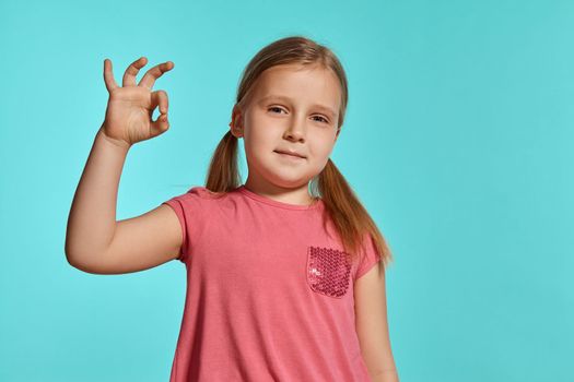 Close-up shot of a pretty blonde little girl with two ponytails on her head, in a pink dress, showing ok sign and smiling against a blue background with copy space. Concept of a joyful childhood.