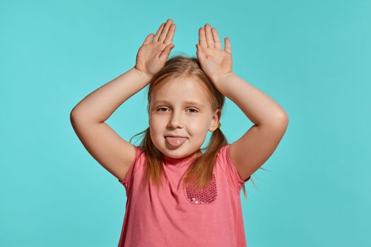 Close-up shot of a beautiful blonde little lady with two ponytails on her head, in a pink dress, making faces and showing her tongue against a blue background with copy space. Concept of a joyful childhood.