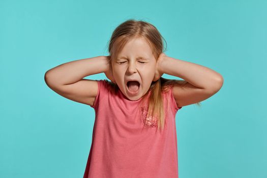 Close-up shot of a charming blonde little girl with two ponytails on her head, in a pink dress, closed her ears with hands and screaming against a blue background with copy space. Concept of a joyful childhood.