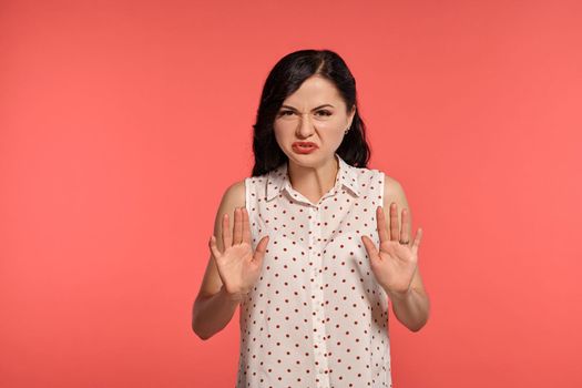 Studio shot of a pretty teeny girl, wearing casual white polka dot blouse. Little brunette femaleis acting like disdaining something and looking at the camera posing over a pink background. Gesticulation and sincere emotions concept.