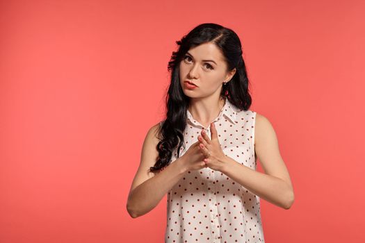 Studio shot of a cute teeny girl, wearing casual white polka dot blouse. Little brunette female is angry, looking at the camera while posing over a pink background. Gesticulation and sincere emotions concept.