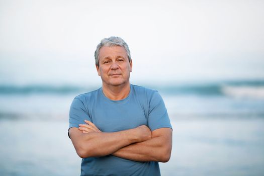 Middle shot of a senior man in blue t-shirt with arms crossed on the chest. Person looking to the camera with a light smile. View against blurry background of sea and sky