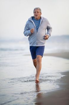 Barefoot senior man in shorts and hoodie jogging on the beach. Coastal sea waves washing his feet. Active and healthy lifestyle in retirement