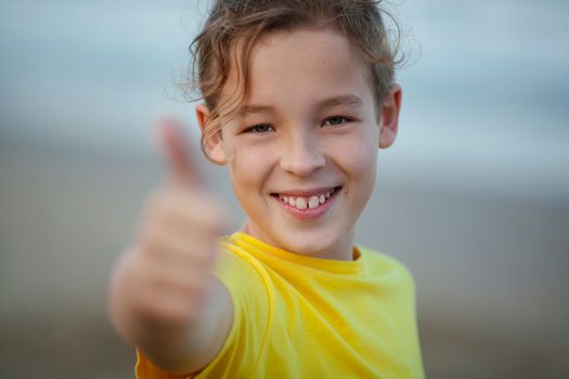 Close-up shot of a happy boy showing thumbs-up. Child with a broad smile in yellow t-shirt on blurry background. Positive thinking