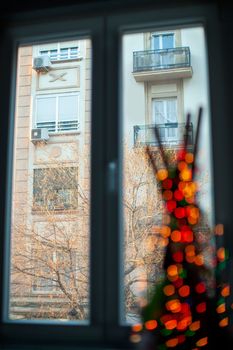 Inside view of a window in the apartment overlooking the neighbouring house and bare tree. Decoration item with blurry red lights in foreground. Focus on the opposite building