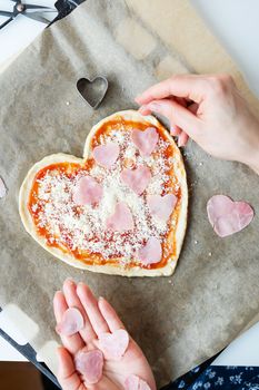 Pizza dough in the shape of a heart, the chef smears the pizza with tomato sauce, sprinkles with Parmesan cheese and decorates it with ham in the shape of a heart. The concept of a surprise for St. Valentine's