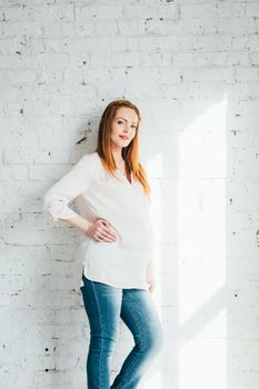 red-haired pregnant girl in a light blouse and blue jeans near the white wall of a large bright room