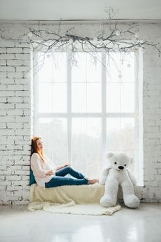 red-haired pregnant girl in a light blouse and blue jeans with a teddy bear on the window