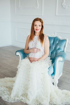 red-haired pregnant young girl in a blue armchair of a bright room