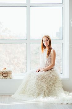 red-haired pregnant young girl in a white dress near the window of a bright room
