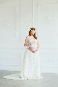 red-haired pregnant young girl in a white dress near the white wall of a large bright room
