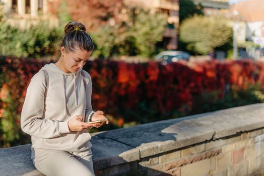 Young fashionable teenage girl with smartphone in park in autumn sitting at smiling. Trendy young woman in fall in park texting. Retouched, vibrant colors. Beautiful blonde teenage girl wearing casual modern autumn outfit sitting in park in autumn. Retouched, vibrant colors, brownish tones.