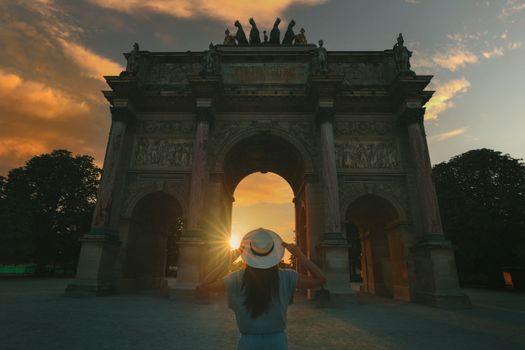 Young woman tourist in sun hat standing in front of Triumphal Arch of the Carousel in Paris at sunset. Travel in France, tourism concept. High quality photo