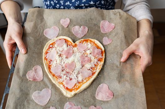 Pizza dough in the shape of a heart, a chef holding a pizza tray decorated with ham in the shape of a heart. Valentine's day surprise concept
