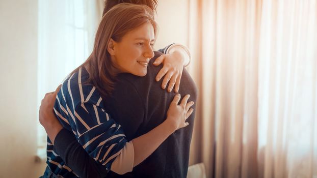 Happy young girl warmly hugs her boyfriend, rejoices at the meeting, stands near the window, smiley face in close-up, woman starts a romantic relationship. Husband and wife feel united, in a harmony.