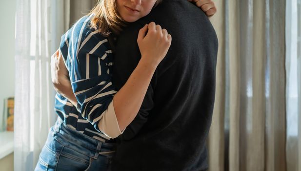 A young girl warmly and tenderly hugs her boyfriend, a couple stands against the background of a window on a sunny day, a woman begins romantic relationship. Husband and wife feel united, in harmony.