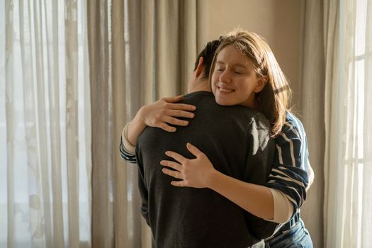 Happy young girl warmly hugs her boyfriend, rejoices at the meeting, stands near the window, smiley face in close-up, woman starts a romantic relationship. Husband and wife feel united, in a harmony.