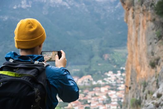 Young man with a backpack and hiking equipment walks in the mountains and takes a photo on the mobile phone of a beautiful picturesque village. Traveler enjoys the scenery and shares photos.