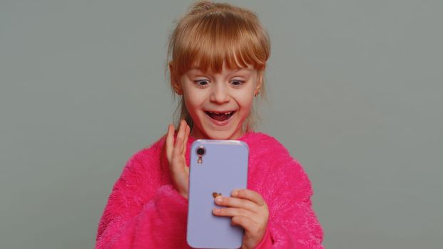 Young preteen child girl kid use mobile smartphone typing browsing say Wow yes found out great big win good news celebrate doing winner gesture. Little toddler children on studio gray wall background