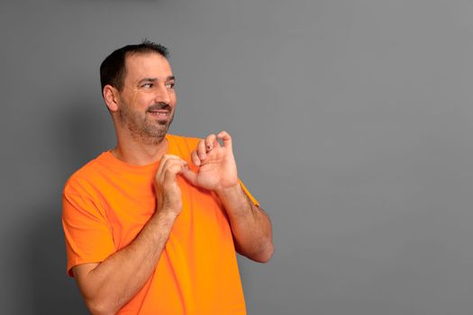 Hispanic man with a beard wearing an orange t-shirt looking to the side with a shy and embarrassed expression. Isolated on gray background