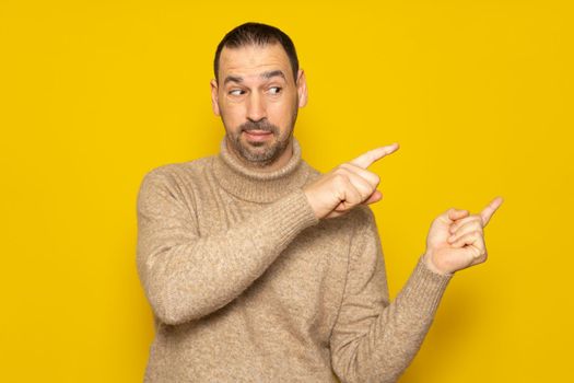 Hispanic man wearing a turtleneck sweater looking at the camera pointing with two hands and fingers to the side, isolated over yellow background.