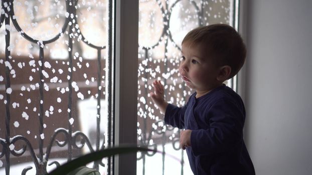 A small child stands on the windowsill and looks out the window. A child knocks on glass in winter. 4k