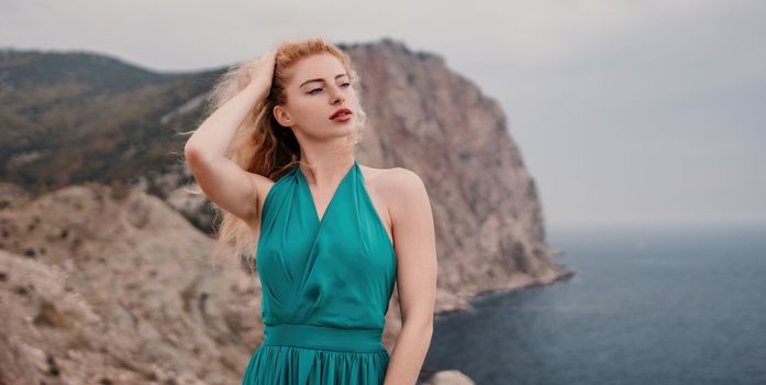 Side view a Young beautiful sensual woman in a mint long dress posing on a volcanic rock high above the sea during sunset. Girl on the nature on overcast sky background. Fashion photo