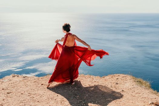 Side view a Young beautiful sensual woman in a red long dress posing on a rock high above the sea during sunrise. Girl on the nature on blue sky background. Fashion photo.