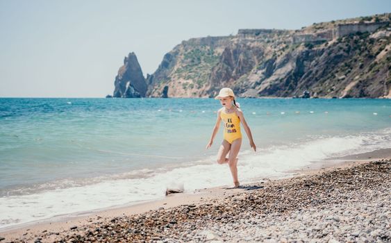 Cute little girl running along the seashore against a clear blue sea and rejoices in the rays of the summer sun. Beautiful girl in yellow swimsuit running and having fun on tropical beach