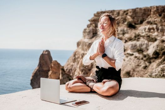 Happy girl doing yoga with laptop working at the beach. beautiful and calm business woman sitting with a laptop in a summer cafe in the lotus position meditating and relaxing. freelance girl remote work beach paradise