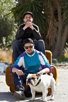 Urban lifestyle. Two gangsters sitting on a couch that is situated in the middle of a street with their dog